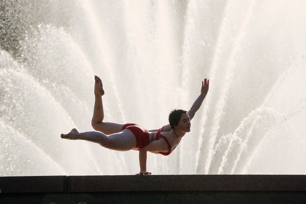 Woman in a red swimsuit performs a one-arm handstand balancing pose outdoors by a large water fountain with water jets, USA.