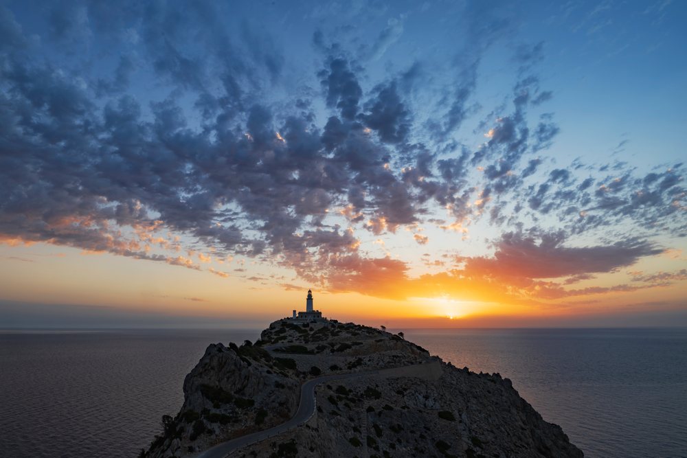 White lighthouse atop rocky cliff at sunrise over the Mediterranean Sea on Cap Formentor, with blue sky and scattered clouds, Landscape, Lifestyle, Pollença, Mallorca, Spain.