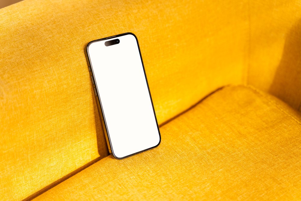 Black smartphone with a blank white screen resting on a yellow fabric sofa in a home interior setting, mock up, Spain.