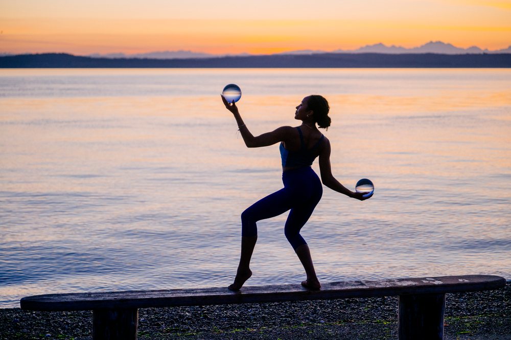 Woman balancing on a bench holding two crystal balls at sunset by the water, yoga, acrobatics, USA.