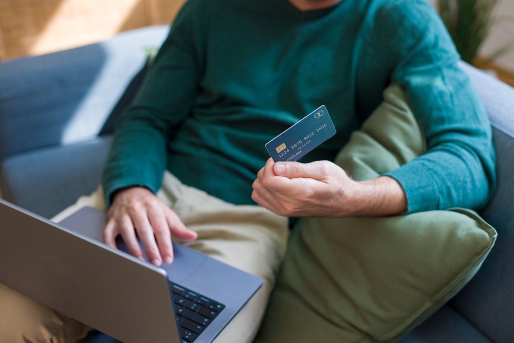 Person sitting on a sofa a credit card and using a laptop indoors, engaging in an online transaction, Technology, young adult, home.