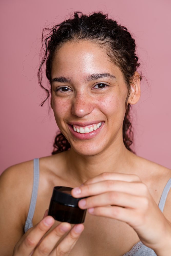 Smiling woman with curly hair holding a cosmetic jar in a beauty shoot against a pink background, Spain.