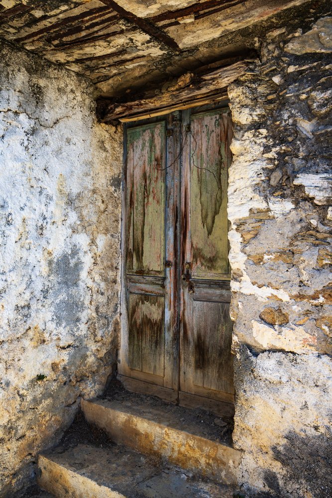 Weathered wooden door with peeling green paint set in a rustic stone wall showing rust stains, Travel Photography, Castellare di Mercurio, Corsica, France.