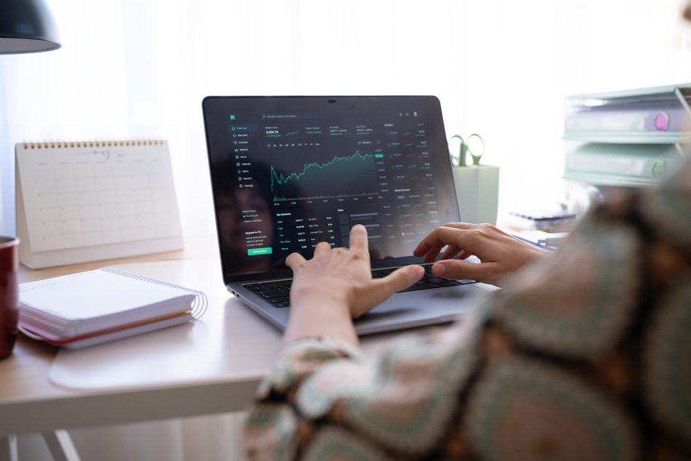 Person typing on a laptop showing stock market data in a bright home office with notebook and paper calendar on desk, Spain.