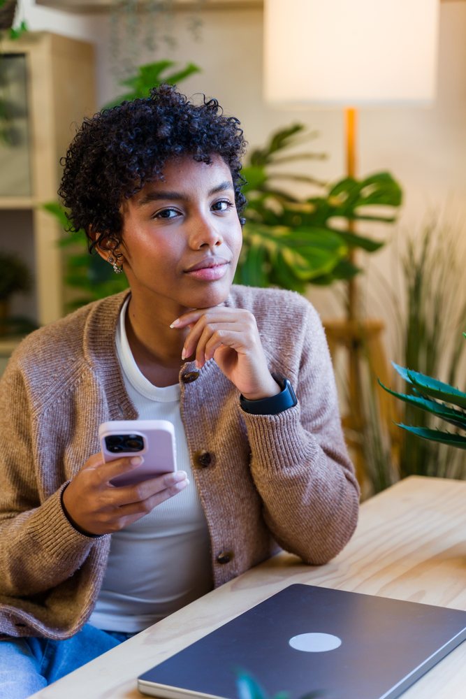 Woman using smartphone at desk with laptop and indoor plants, Spain.