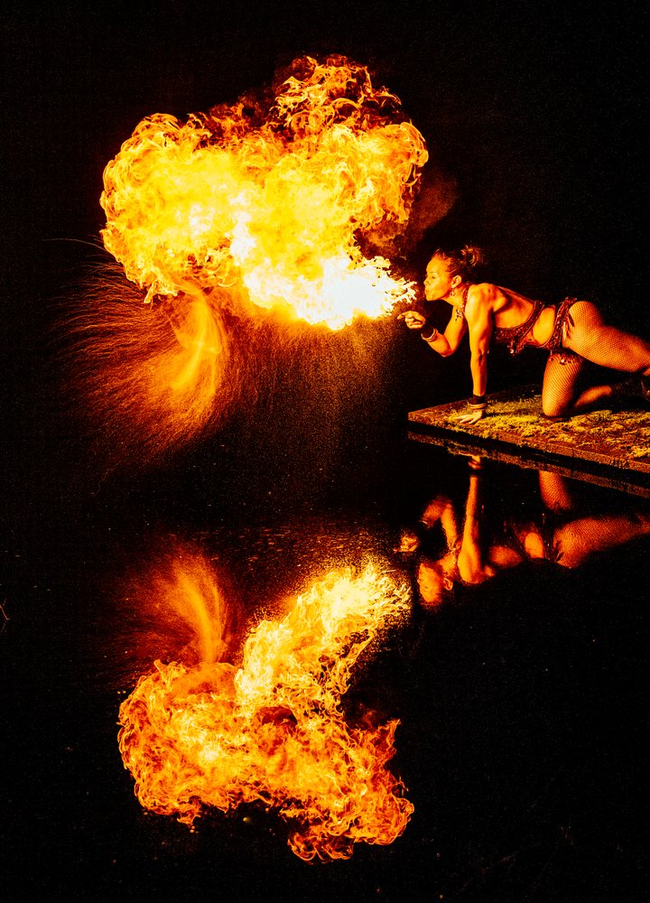 Muscular woman performing fire breathing at night with flames and her reflection visible on the water surface, artistry, artist, USA.