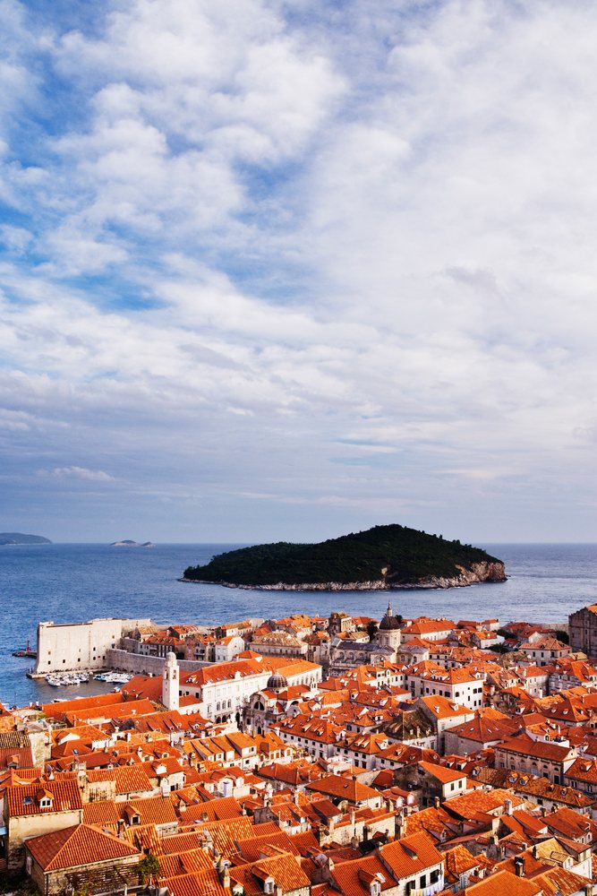View of the Old City of Dubrovnik with red rooftops and stone fortress walls overlooking Lokrum Island and the Adriatic Sea under a cloudy sky.