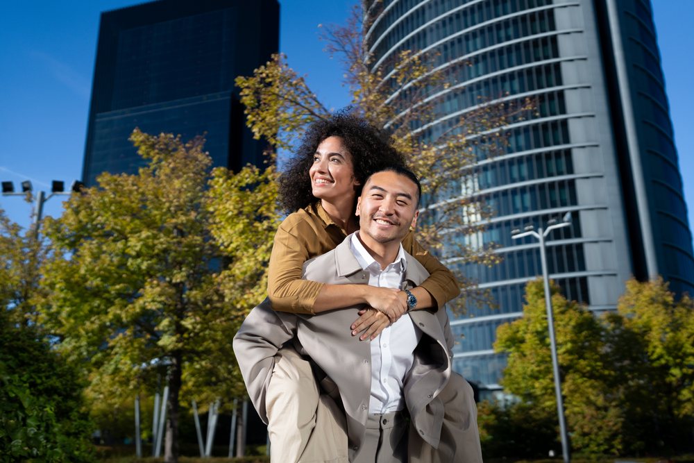 Man giving woman a piggyback ride outdoors in an urban park with modern buildings and trees under blue sky, Madrid, Spain.