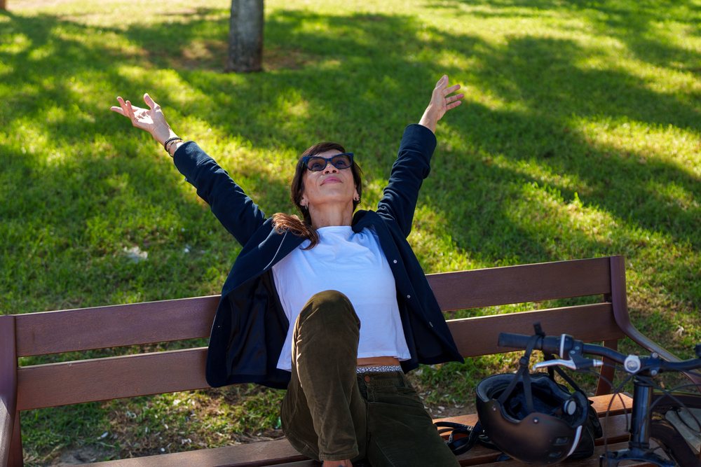 Woman sitting on a wooden bench stretching her arms outdoors beside a parked bicycle and helmet in a sunny park, Málaga.