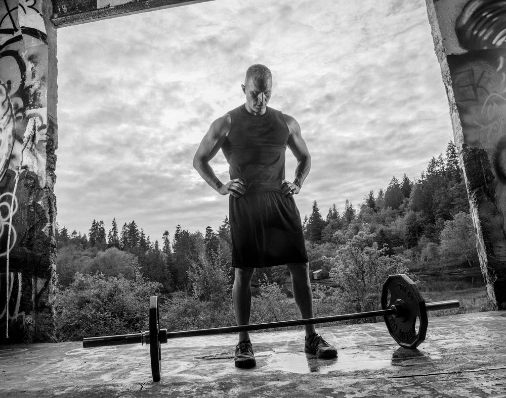 Muscular man stands with hands on hips outdoors near an abandoned building preparing to lift a barbell with weights on the ground, forest visible behind, USA.