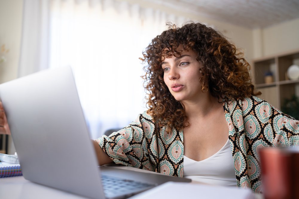 Woman with curly hair uses a laptop for a video call while seated at a table in a home office, Spain.