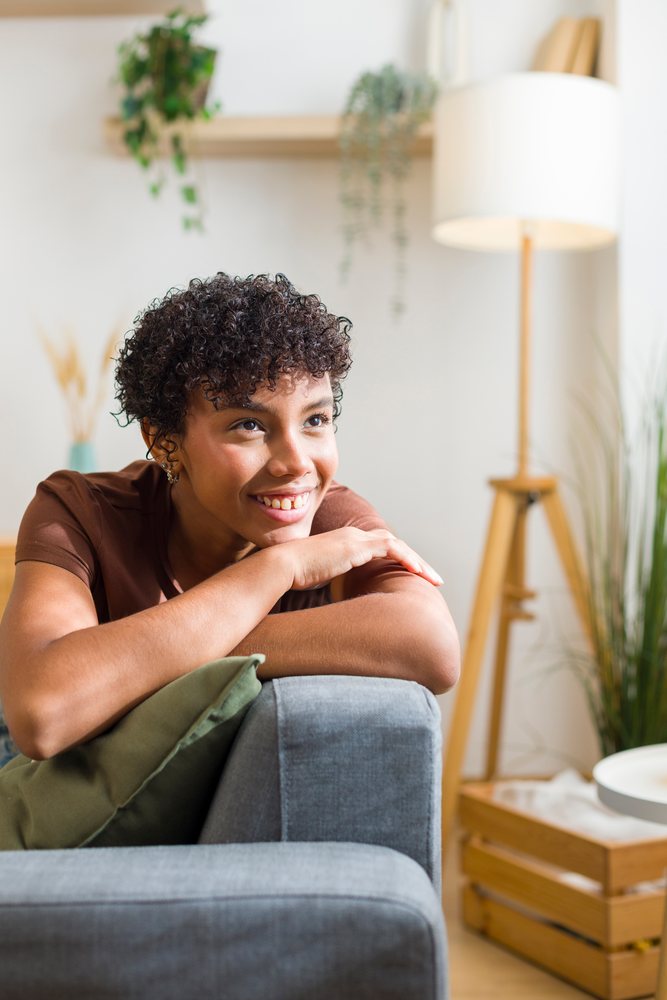 Young woman smiling and leaning on the armrest of a grey couch in a cozy living room with houseplants and a floor lamp.