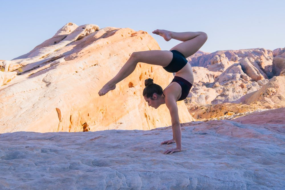 Woman performing a handstand yoga pose on rocky desert terrain in the USA with large rock formations in sunlight.