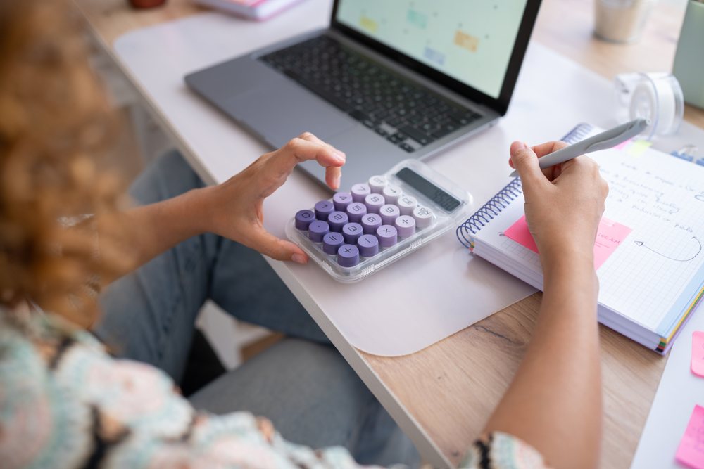 Woman using calculator and writing notes in a notebook at a desk with a laptop in a home office, Spain.