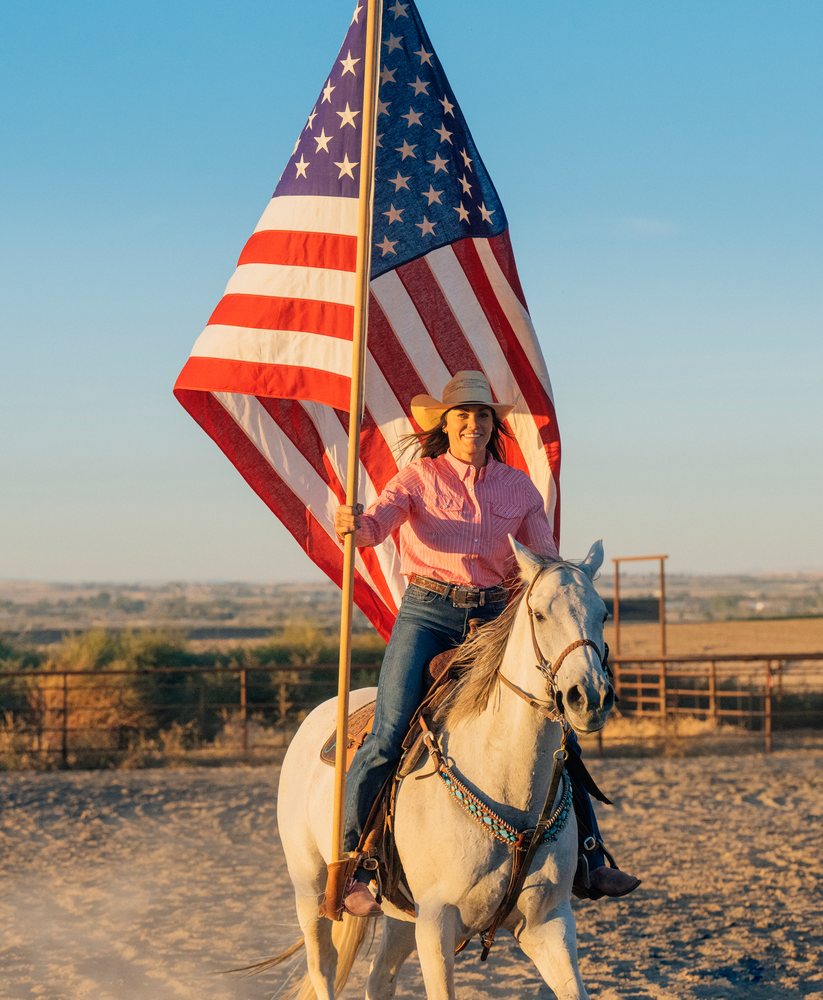 Woman riding a white horse holding an American flag in a dusty outdoor corral at sunset, Southwest USA.