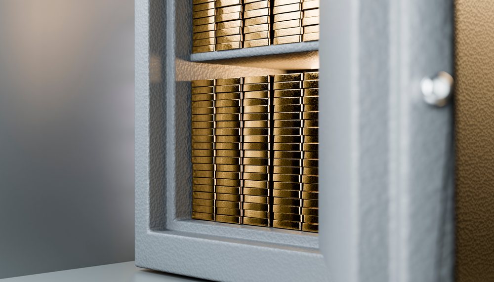 Stacked gold bars with shiny metallic surface inside a safety deposit box with a partly open gray metal door.