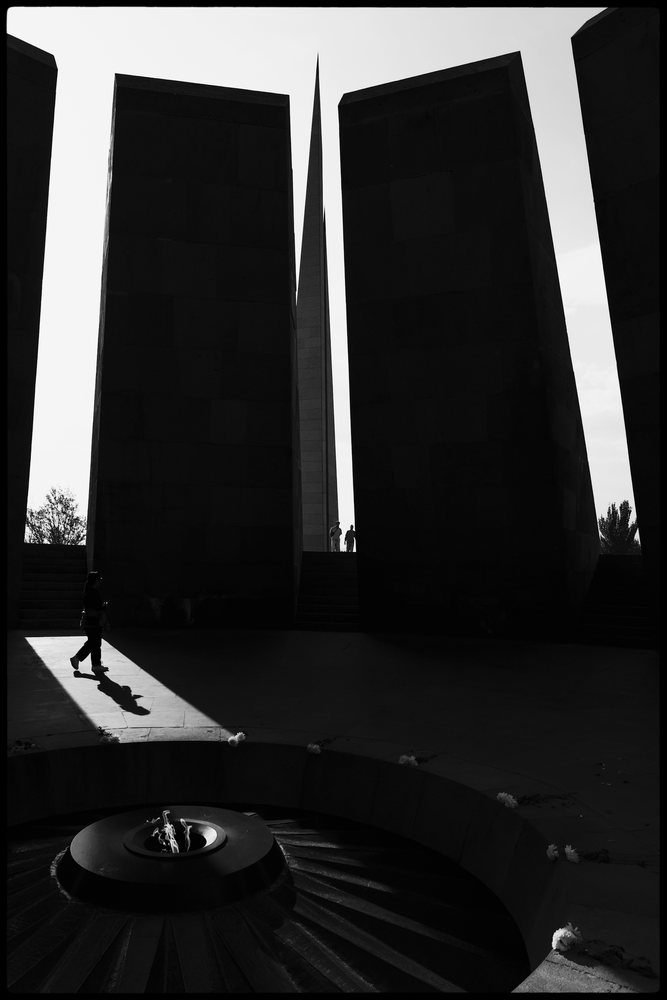 The Armenian Genocide Memorial, features large stone spires, a circular stone structure with a central flame, and visitors walking near the monument, Yerevan, Armenia.
