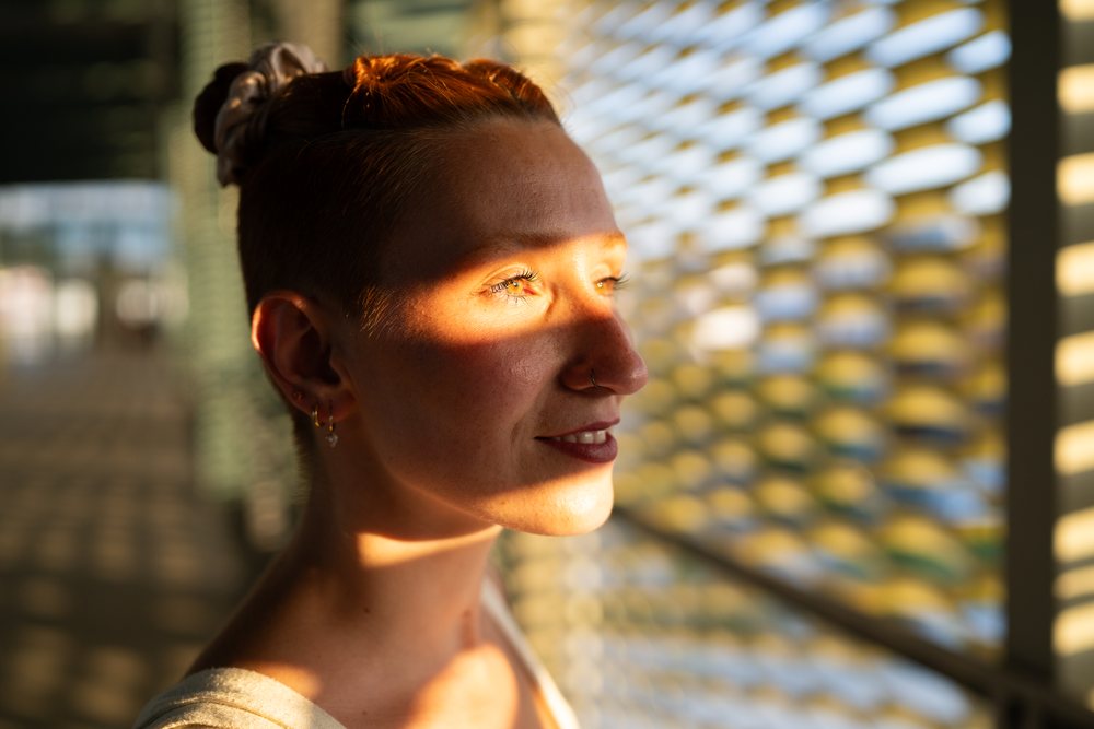 A young woman with earrings and a nose ring smiles softly as sunlight creates a dynamic shadow pattern across her face through window blinds indoors, urban, Santiago De Compostela, Spain.