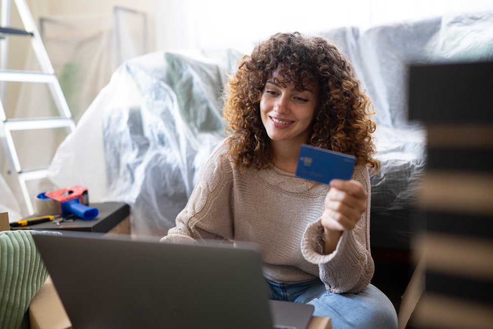 Woman sitting on the floor indoors using a laptop and holding a credit card amid moving boxes and plastic-covered furniture, Spain.