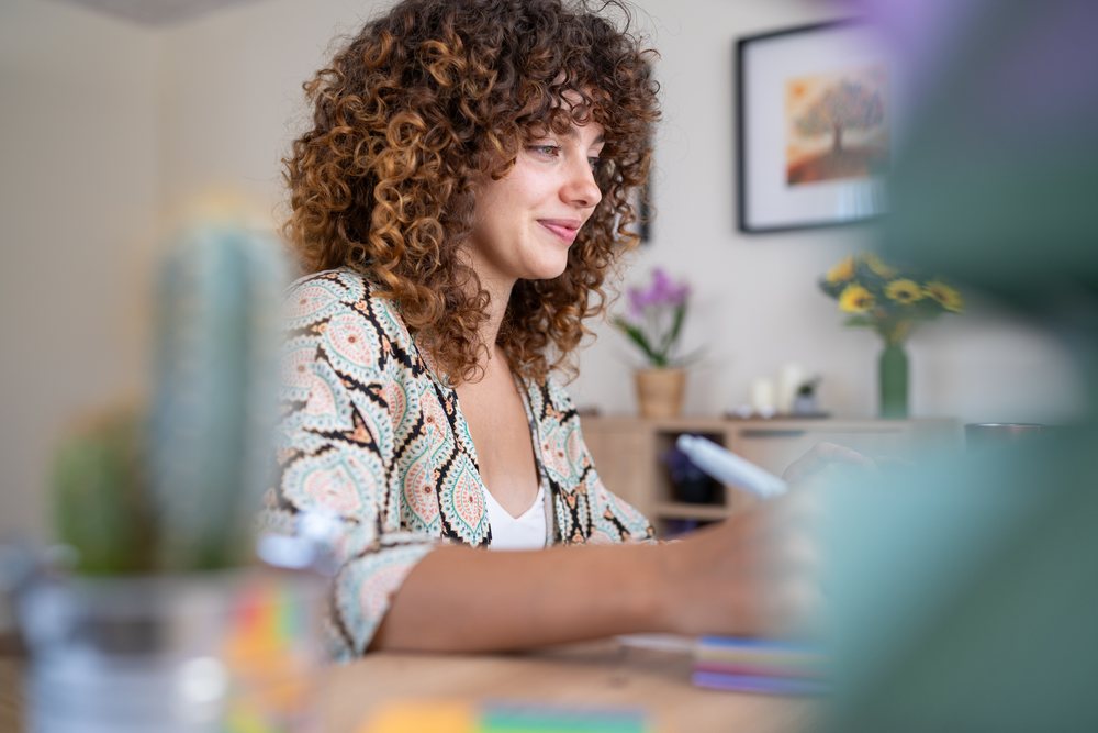 Woman with curly hair writes at a desk in a Spanish home interior decorated with plants and artwork, Spain.
