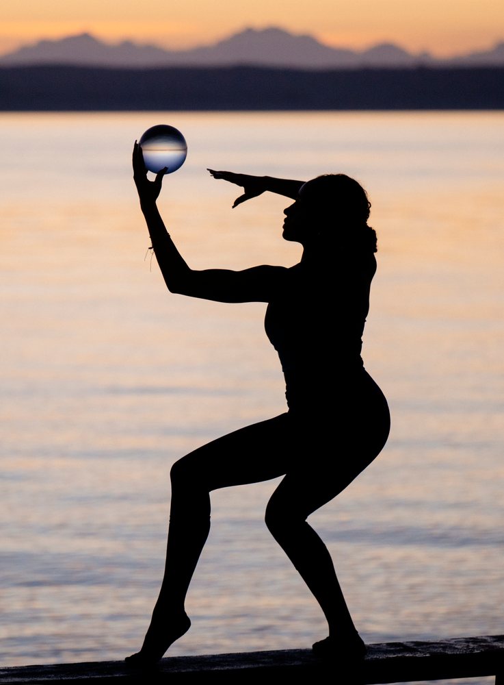 Silhouette of a woman performing an acrobatic yoga pose holding a glass ball at sunset by the water, USA.