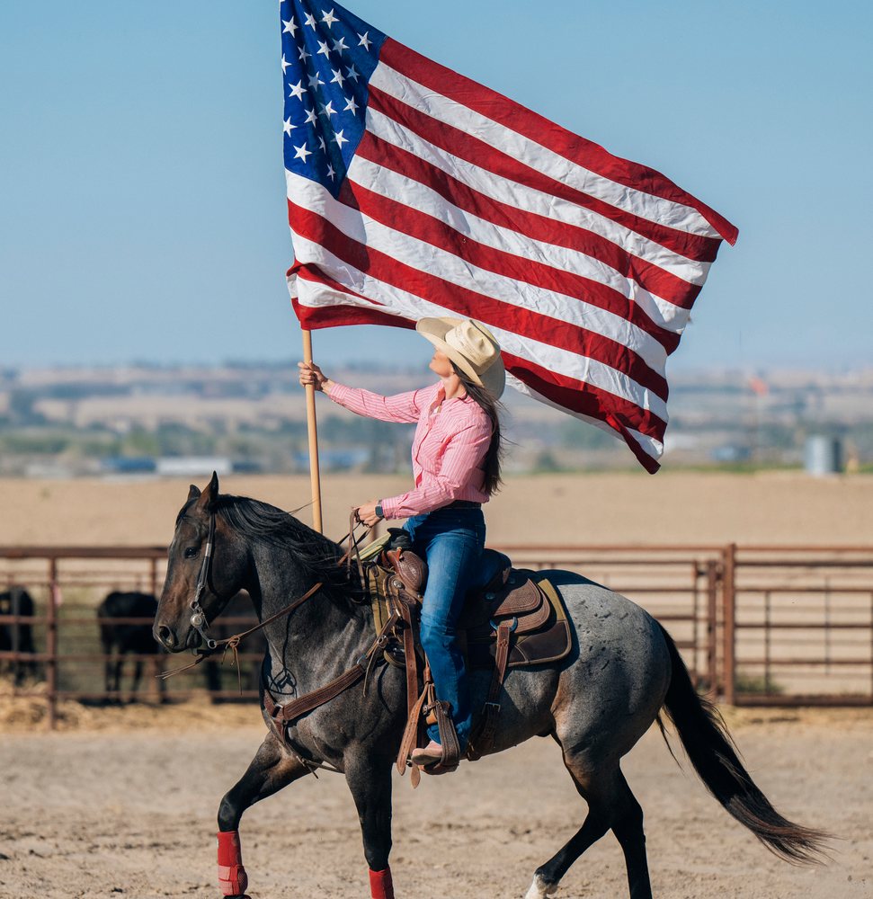 Woman wearing a cowboy hat rides a male horse holding a large American flag at a Southwest USA ranch outdoors under a blue sky.