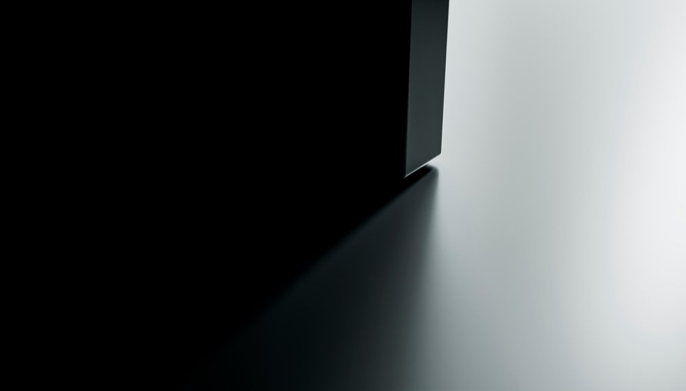 Shadow cast by the sharp corner of a black rectangular object falls on a smooth white surface creating high contrast in a minimalist studio setting.
