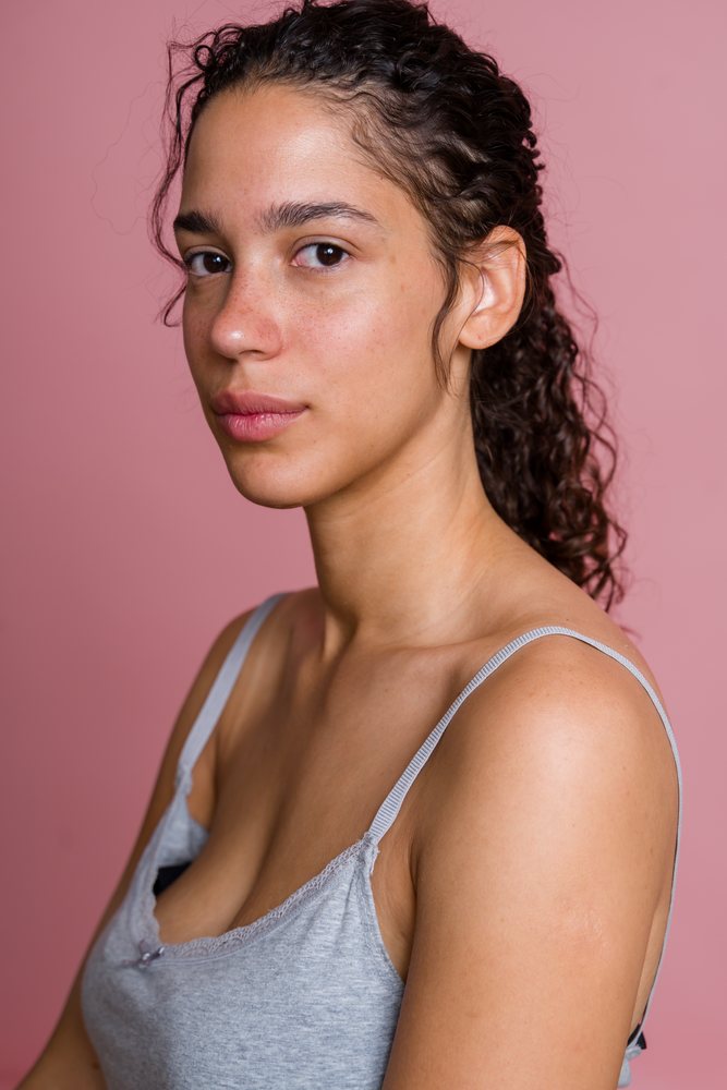 Young woman with curly hair wearing a grey tank top poses against a pink background in a studio portrait, beauty shoot, Spain.