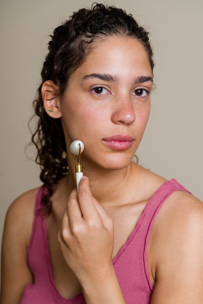 Woman with curly hair uses a jade roller for facial massage in a beauty studio setting, Spain.