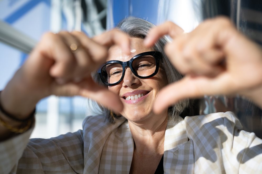 Smiling mature woman with gray hair and black eyeglasses making a heart shape with her hands indoors, businesswoman, traveler, Spain, Madrid.