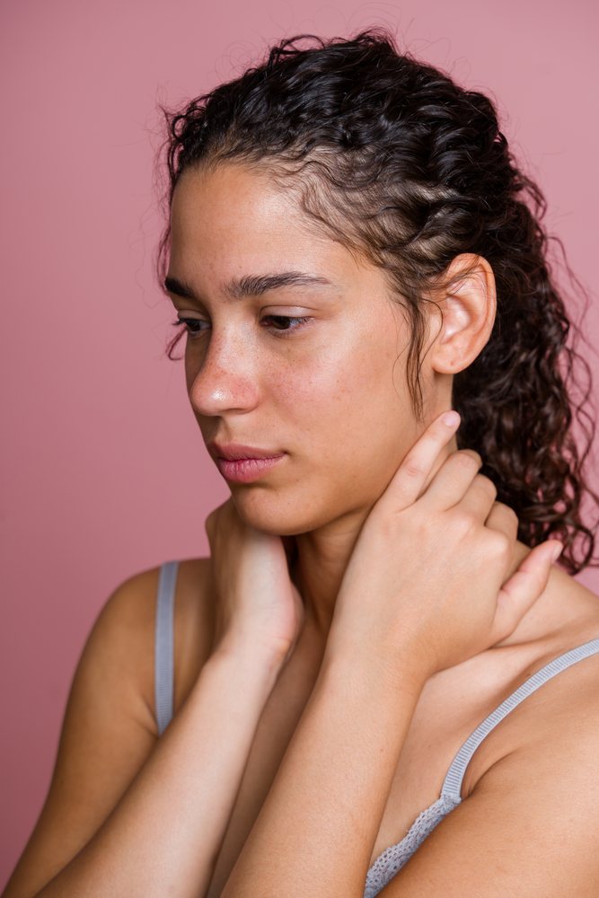 Woman with curly hair wearing a grey tank top holds her neck against a pink background in an indoor beauty shoot, Spain.