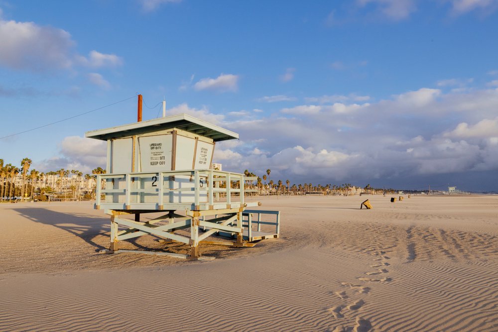 Lifeguard tower centered on a sandy Venice Beach shore with palm trees and partly cloudy blue sky in the background, CA, USA.