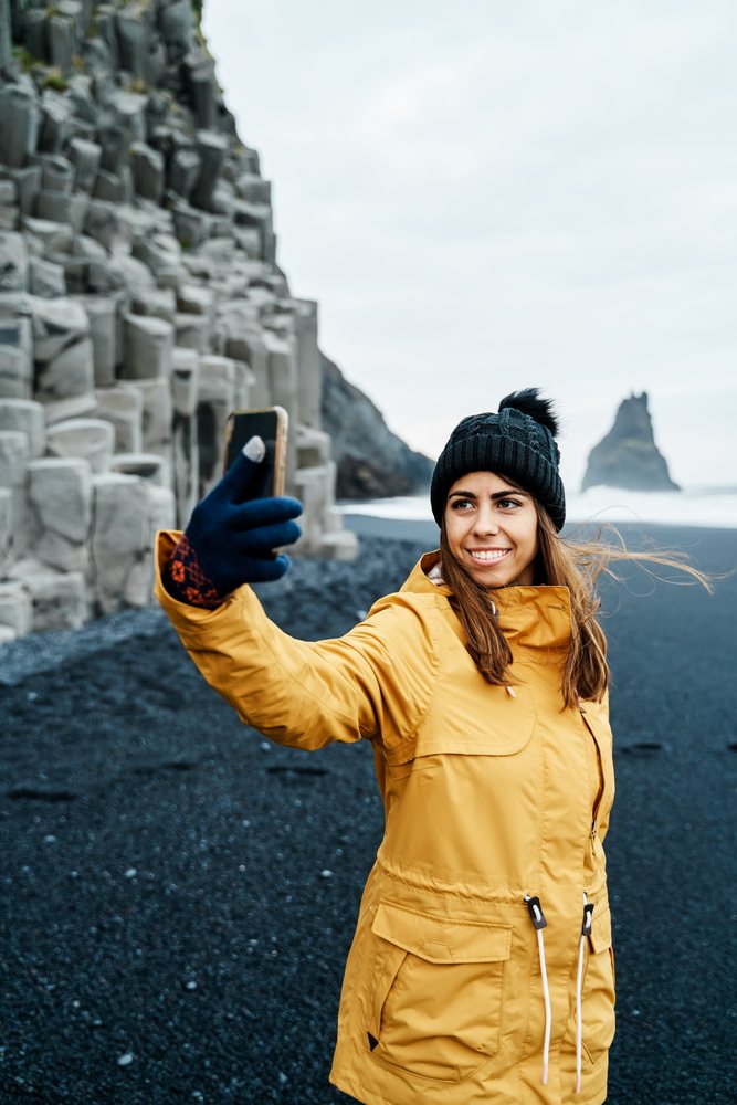 A white woman takes a selfie on a black sand beach in a beach