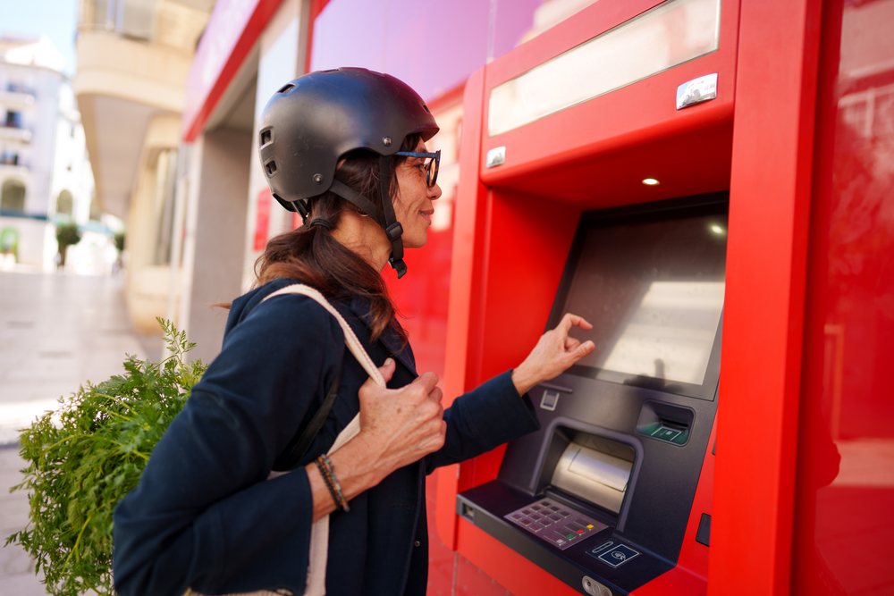 Woman wearing a black helmet and glasses uses a red ATM outdoors on a street in Málaga while carrying a shopping bag with fresh carrots.