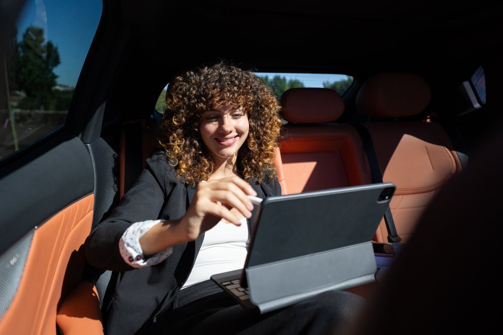 Woman with curly hair sitting in car back seat using a tablet computer for remote work in Spain during daylight.