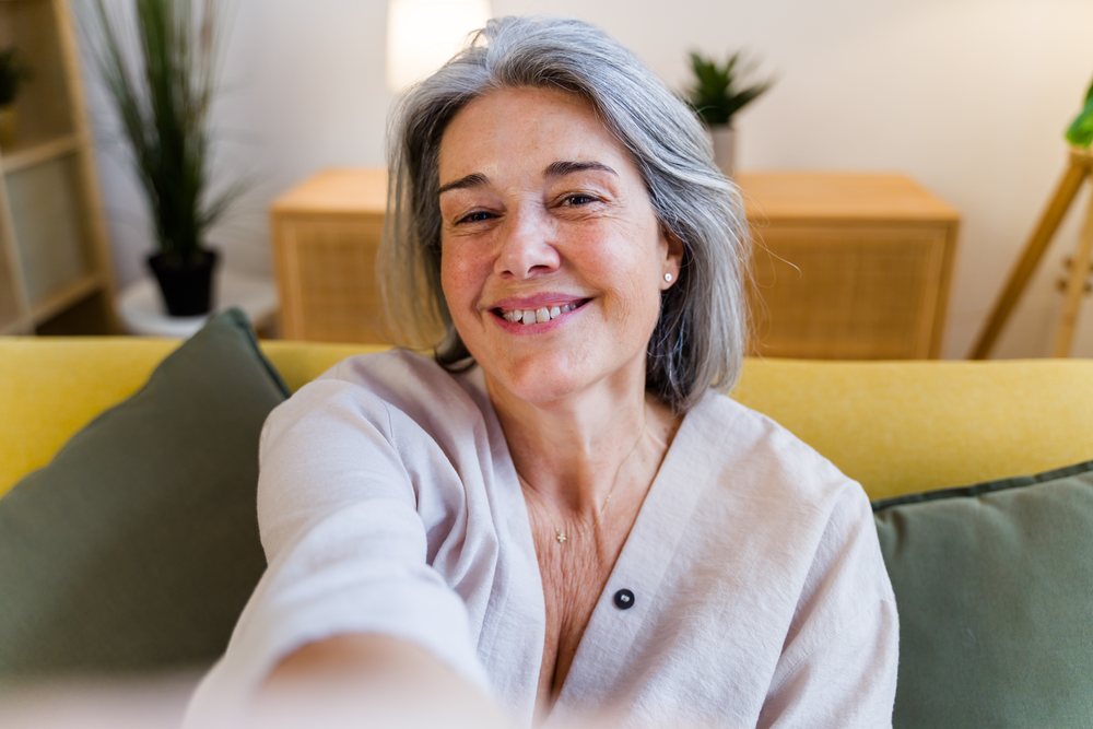 Middle-aged woman with gray hair smiles while taking a selfie seated on a couch indoors with a pillow and plants nearby, Spain.