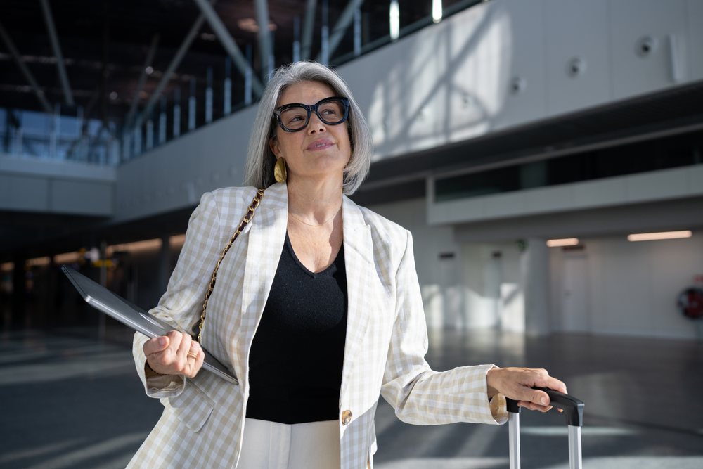 Mature businesswoman with gray hair and glasses stands holding a laptop and suitcase inside a modern airport terminal, Spain, Madrid.