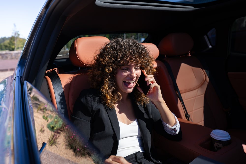 Woman with curly hair wearing a black blazer talks on phone while sitting inside a car with leather seats and a coffee cup in the holder, remote work, businesswoman, Spain.