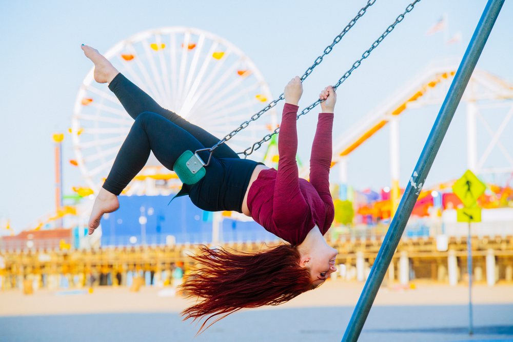 Barefoot woman with red hair swings upside down on a swing at an amusement park near a pier and ferris wheel during daytime, California, USA.