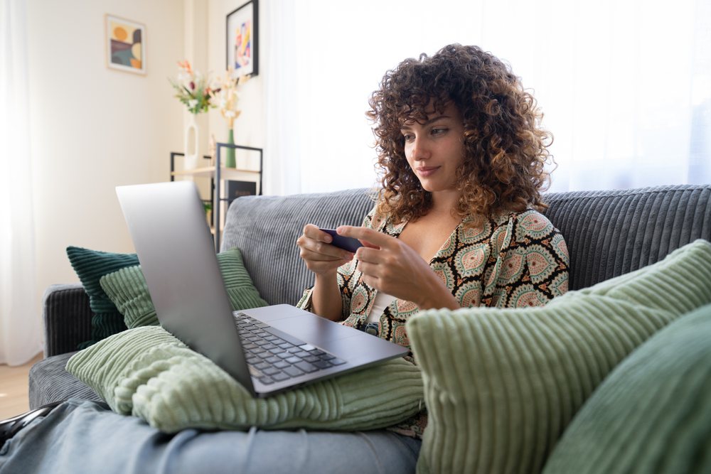 Woman with curly hair sits on a gray couch using her smartphone, resting a laptop on green pillows in a sunny living room with decorative elements, home, girl, Spain.