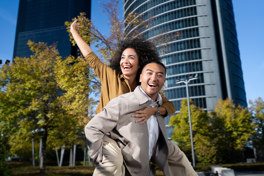 Couple enjoying a piggyback ride outdoors in an urban park with skyscrapers and trees in the background, Madrid, Spain.