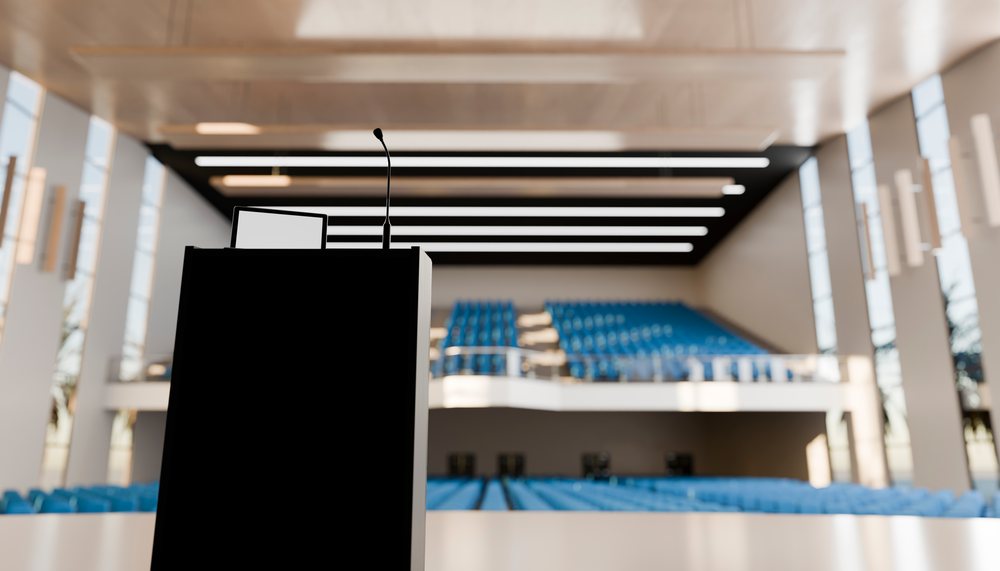 Podium with microphone and screen in front of rows of blue seats in an empty lecture hall auditorium.