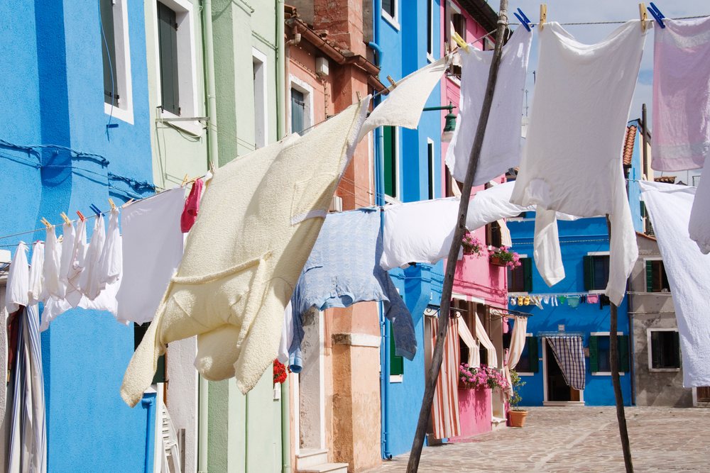 White and light-colored laundry hangs on multiple clotheslines across a narrow colorful residential street on a sunny day, Venice, Italy.