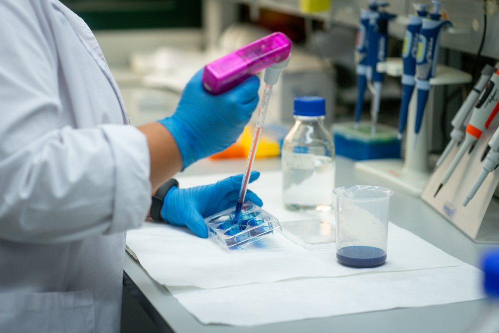 Scientist wearing blue gloves and white coat uses a pipette to transfer blue liquid into a plastic container in a Seville research laboratory, Andalucía.