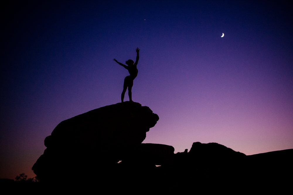 Silhouette of a person performing yoga with uplifted arms on a rock at dusk under a crescent moon, USA.