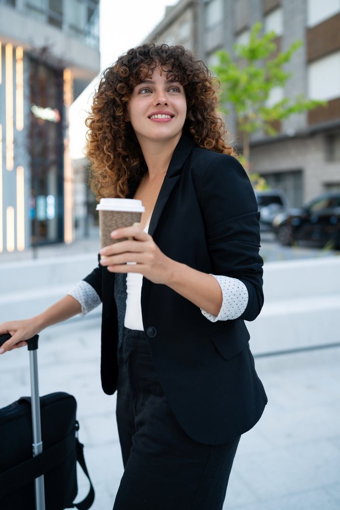 Young woman in business attire holding a coffee cup and pulling a suitcase on an urban street in Madrid during daytime.