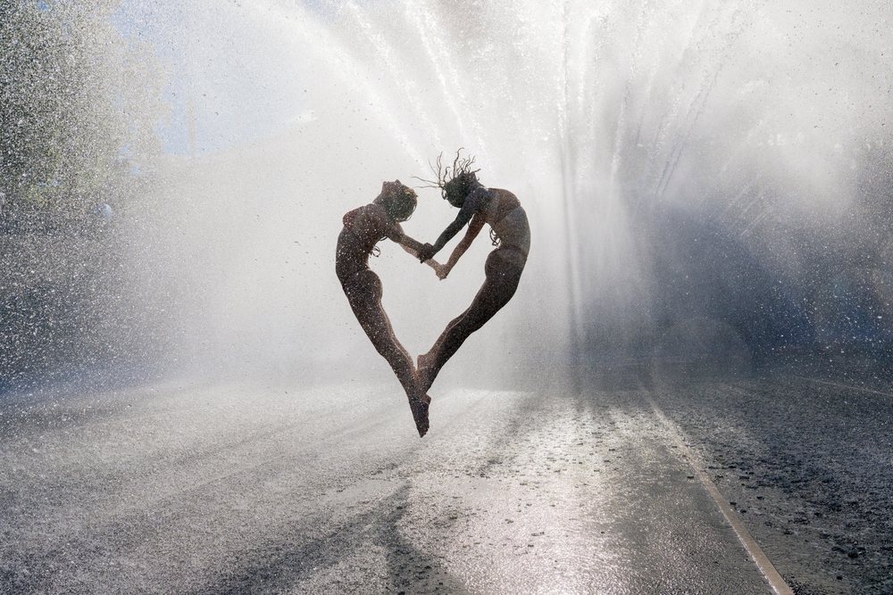 Two women jumping and holding hands in a heart shape while surrounded by water spray from a sprinkler outdoors, partner, acrobatics, yoga, USA.
