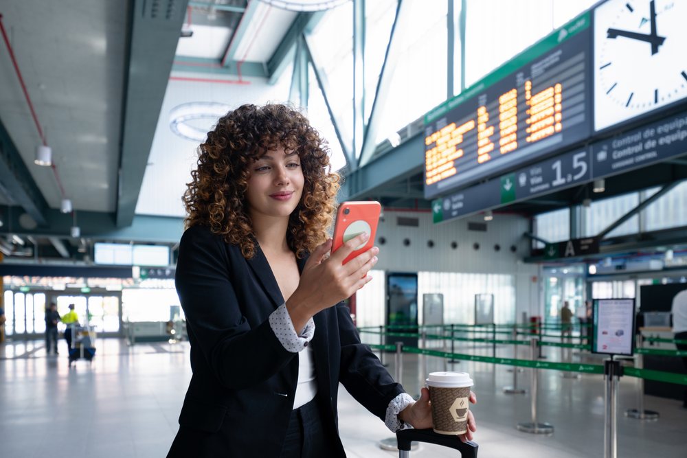 Woman with curly hair stands in an airport terminal holding a coffee cup and using a smartphone near a departure board and luggage, Spain.