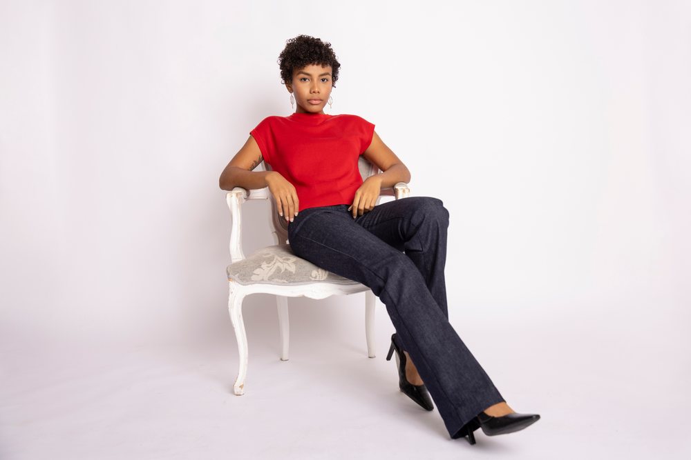 Woman with curly hair sits on a white chair wearing a red top, black pants, and black high heels in a studio setting, fashion, clothes, young culture.
