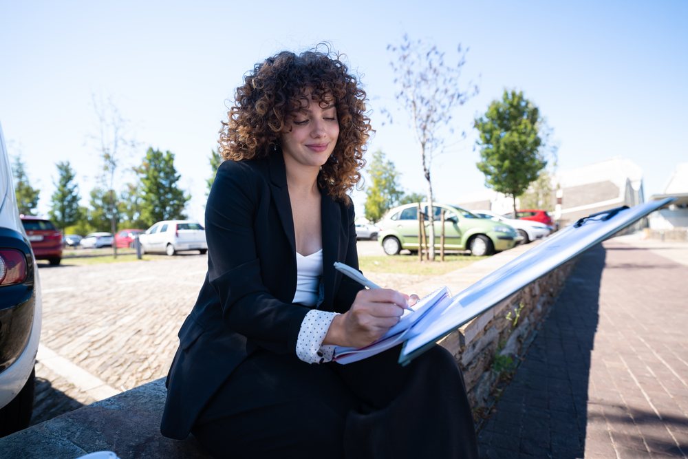 Businesswoman wearing a black suit is writing notes outdoors while seated in an urban car park area on a sunny day, traveling executive, Spain.